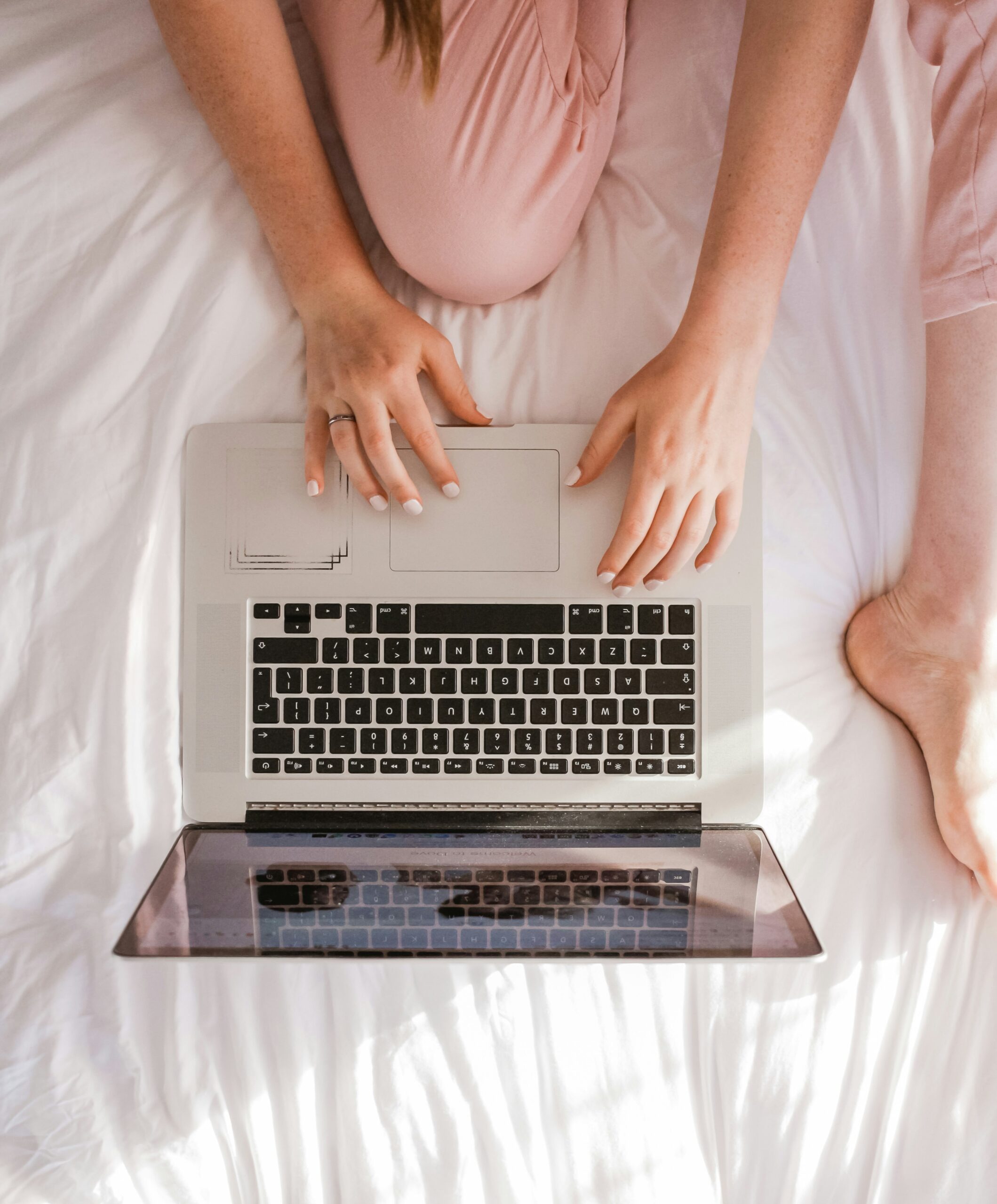 A woman in pink pajama pants uses a laptop in bed to attend virtual neurodiversity-affirming therapy.