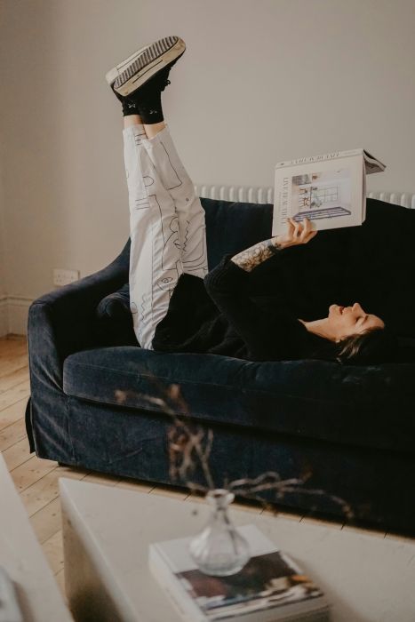 A person lies on a dark couch reading a book with their feet straight up in the air, considering an adult AuDHD assessment.