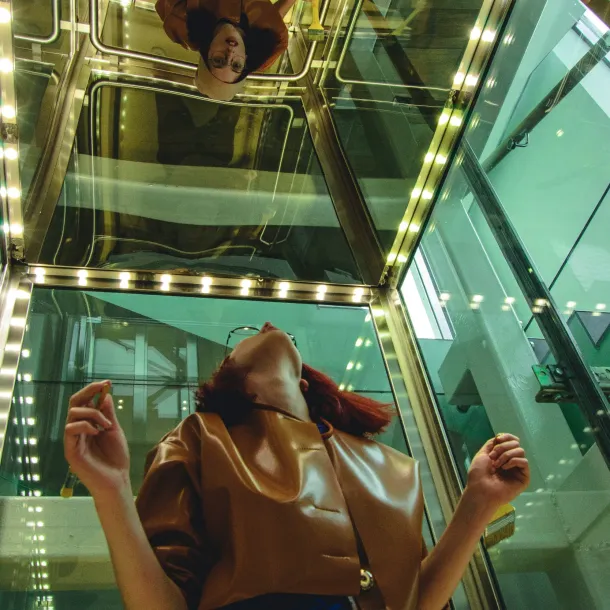 A woman wearing glasses gazes upward in a glass elevator, as if looking for adult autism resources.