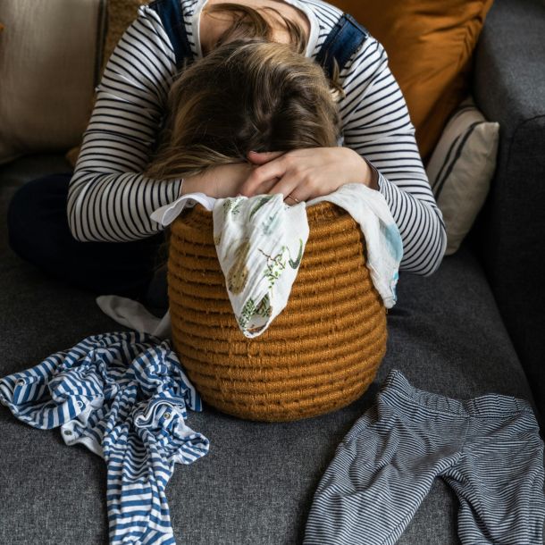 A woman sits on a couch hiding her face with her hands, reflecting feelings of stress and the challenges of pathological demand avoidance (PDA).