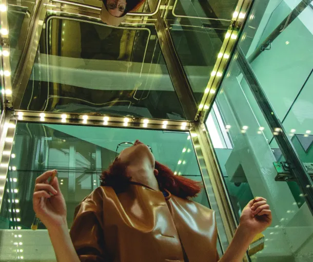 A woman wearing glasses gazes upward in a glass elevator, as if looking for adult autism resources.