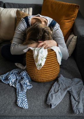 A woman sits on a couch hiding her face with her hands, reflecting feelings of stress and the challenges of pathological demand avoidance (PDA).