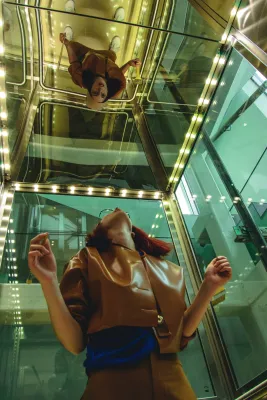 A woman wearing glasses gazes upward in a glass elevator, as if looking for adult autism resources.