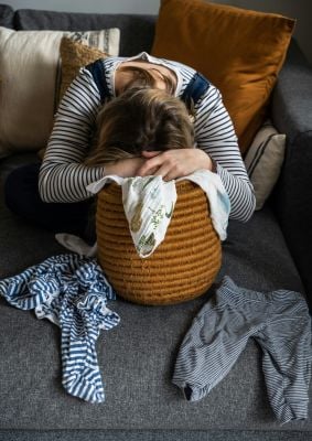 A woman sits on a couch hiding her face with her hands, reflecting feelings of stress and the challenges of pathological demand avoidance (PDA).