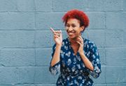 A smiling woman with bright hair stands in front of a blue wall pointing her finger and celebrating a late in life autism diagnosis.